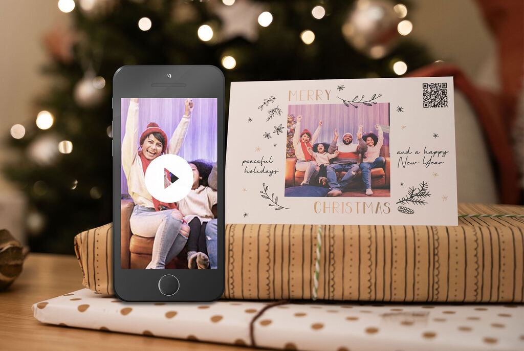 Een persoon houdt een telefoon met een video vast, terwijl een ander op de bank zit en geniet, omringd door cadeaupapier. Op de achtergrond straalt een kerstboom.